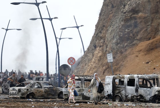Des femmes sur la route de Ceuta, avec en arrière-plan des véhicules brûlés, le 31 juillet 2026 près de la frontière entre l'Espagne et le Maroc.